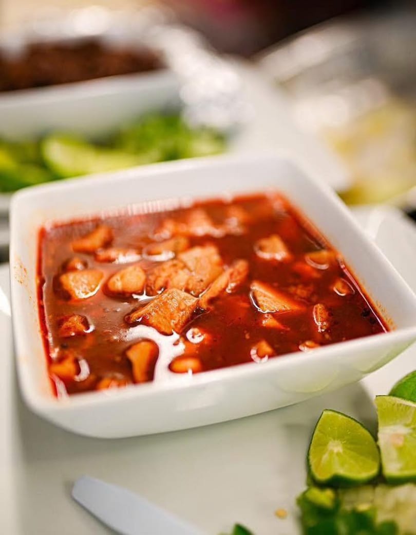 A white bowl of red stew with meat chunks, accompanied by lime wedges and green chilies.
